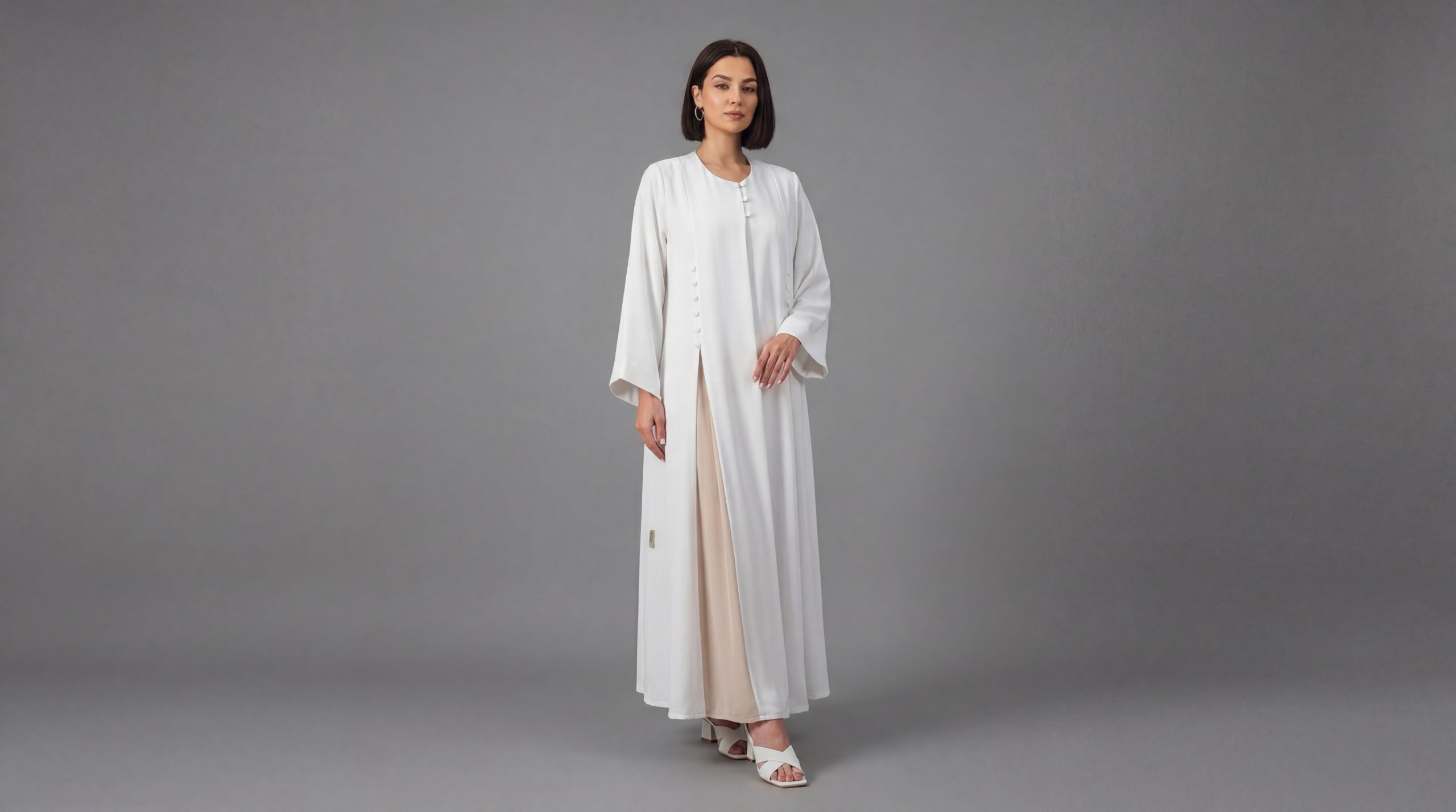 Pure Sophistication: The Ivory Asymmetric Abaya Embrace a new level of minimalist luxury with our Ivory Classic Abaya. This piece stands out with its clean architectural lines and unique asymmetric button fastening that adds a contemporary twist to the classic silhouette. Designed for the woman who loves "Quiet Luxury," it offers a bright, refreshing aesthetic perfect for daytime events, formal gatherings, or professional settings. Key Features: Unique Asymmetric Fastening: Features a row of elegant, fabric-covered buttons placed off-center, creating a modern wrap-effect that is both stylish and modest. Refined Side Slit: A subtle high-positioned slit allows for graceful movement and a glimpse of the coordinating inner dress, adding dimension to the outfit. Premium Ivory Fabric: Crafted from high-quality, opaque crepe with a soft matte finish that feels luxurious against the skin and stays crisp all day. Versatile Layering: The pristine white tone pairs beautifully with earth-toned inners (as pictured) or monochromatic whites for a bold, ethereal look. Modest Round Neckline: A classic, clean neckline that provides full coverage while remaining effortlessly chic. Embrace a new level of minimalist luxury with our Ivory Classic Abaya. This piece stands out with its clean architectural lines and unique asymmetric button fastening that adds a contemporary twist to the classic silhouette. Designed for the woman who loves "Quiet Luxury," it offers a bright, refreshing aesthetic perfect for daytime events, formal gatherings, or professional settings.
